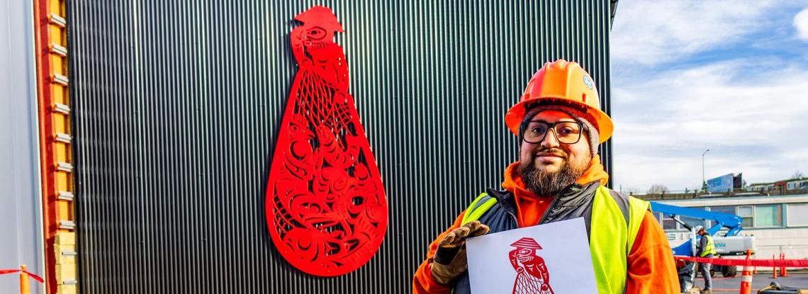 Ty Juvinel poses in the foreground with his piece "Beach Seining" on the Maritime Innovation Center building.