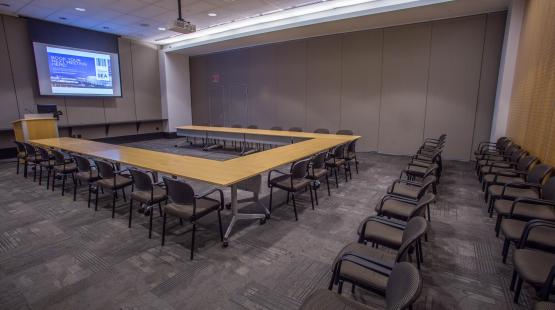 View from the entry doorway looking into the Beijing Conference room that is in a u-shape room set and two rows of theater style seating behind it. The projector is turned and the projector screen is displaying content